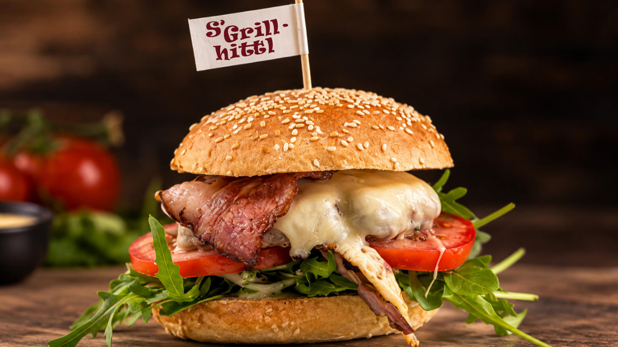 Bacon-Cheeseburger auf einem Sesambrötchen mit einem S'Grillhittl-Fähnchen als Garnierung. - S'Grillhittl